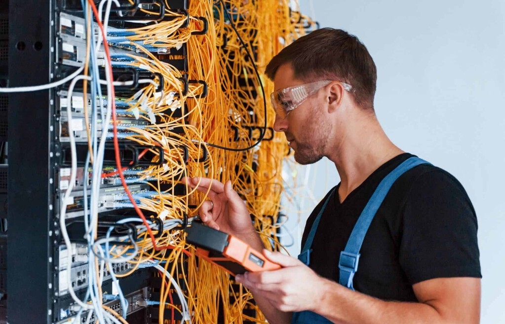 Junger Mann mit Schutzbrille arbeitet mit Internetgeräten und Kabeln im Serverraum